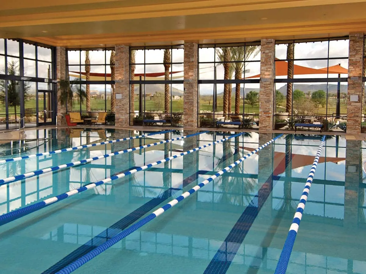An inviting indoor swimming pool area surrounded by large windows revealing palm trees, with ample seating for socializing and relaxing. The pool is bright and lively, ready for fitness training or casual swims.