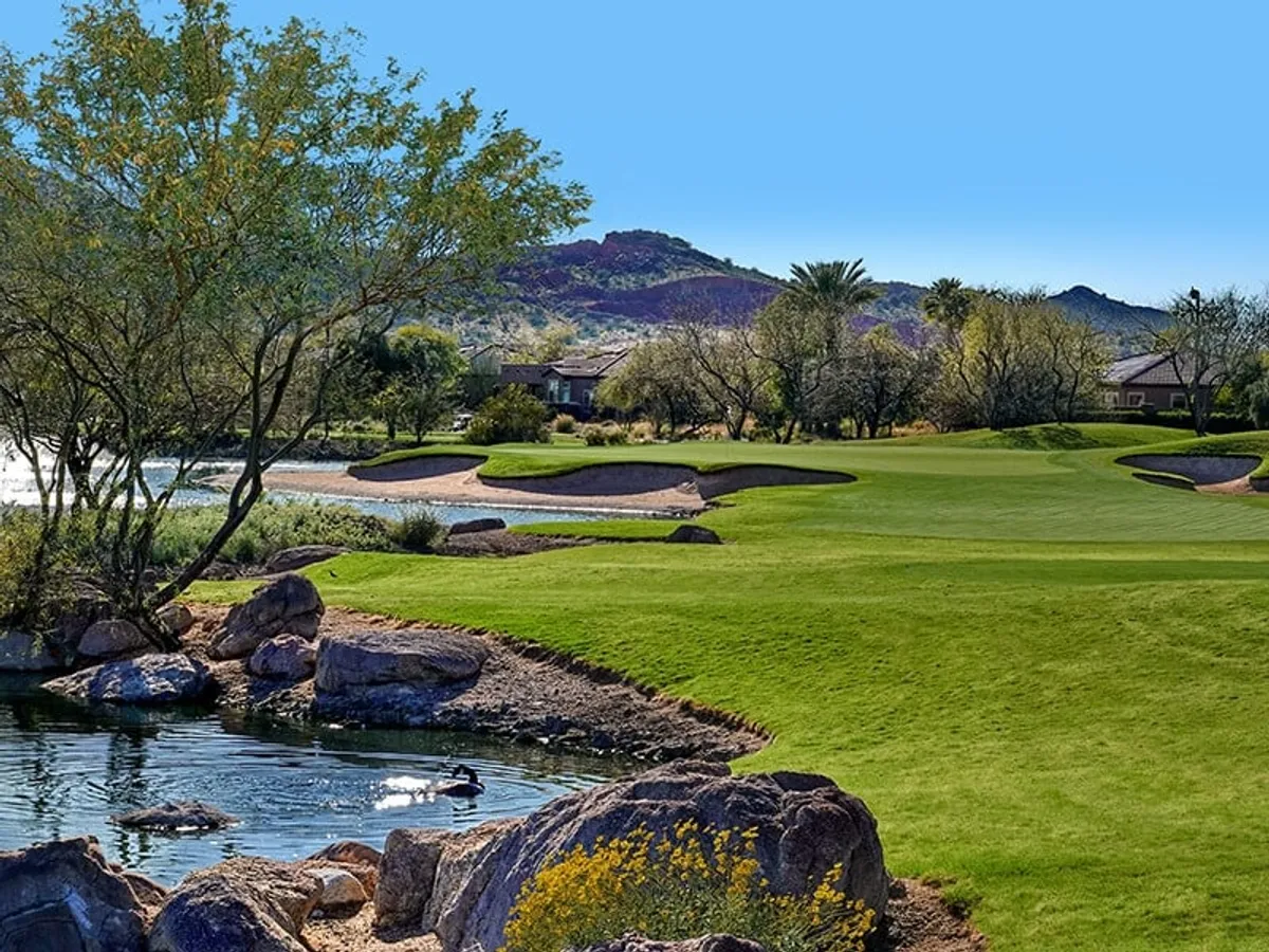 A sweeping view of the Trilogy Golf Club’s lush, manicured fairways bordered by a tranquil lake, mature trees, and distant mountains under a soft sky. The scene captures golfers enjoying a round on the expansive green, with natural water features and desert flora enhancing the picturesque setting.