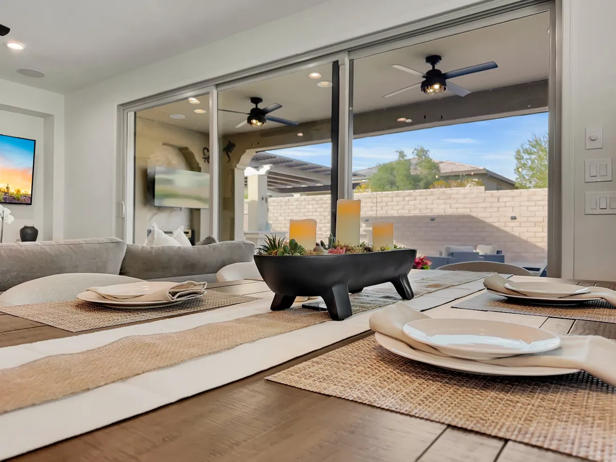 A warmly lit dining area featuring a table set with plates and a decorative centerpiece. Large windows bring in natural light and offer an outdoor view, suggesting a seamless flow between indoor dining and the patio beyond.