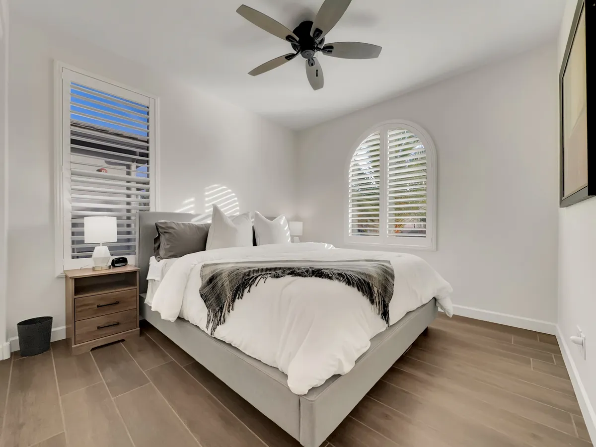 A serene bedroom featuring a king-sized bed with crisp white linens, an arched window that lets in abundant sunlight, and a ceiling fan above. The atmosphere is tranquil, perfect for restful nights or peaceful mornings. Minimalist decor ensures the focus remains on comfort and relaxation.
