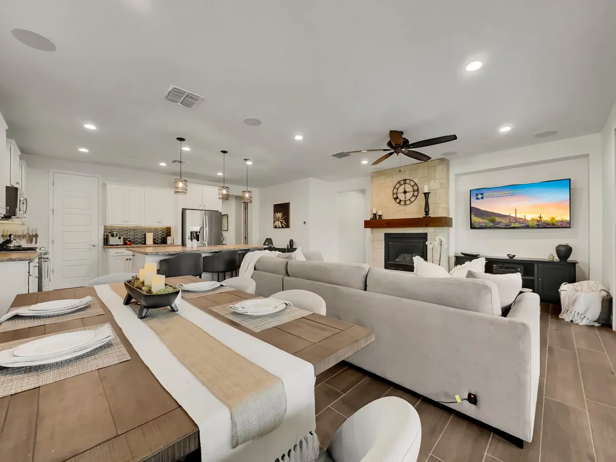 A spacious interior shot featuring a dining table set for a meal, a cushioned sofa, a cozy fireplace with a decorative clock, and a television displaying a scenic image. The arrangement encourages both social dining and casual lounging.