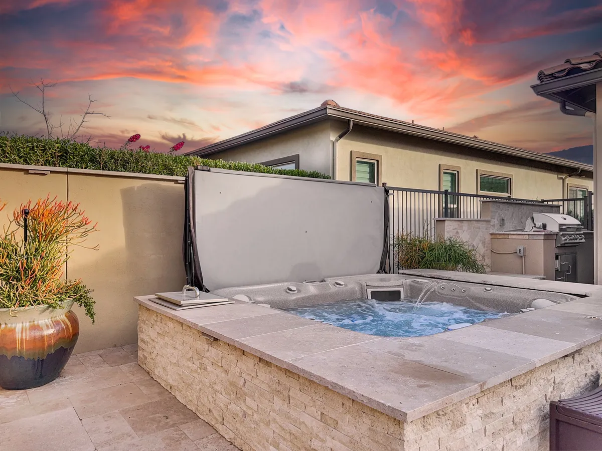 A tranquil garden patio with a bubbling hot tub set amidst manicured landscaping, looking out toward a glowing sunset. The area includes comfortable seating and is designed for both relaxation and social connection, perfect for enjoying Arizona's beautiful evenings.