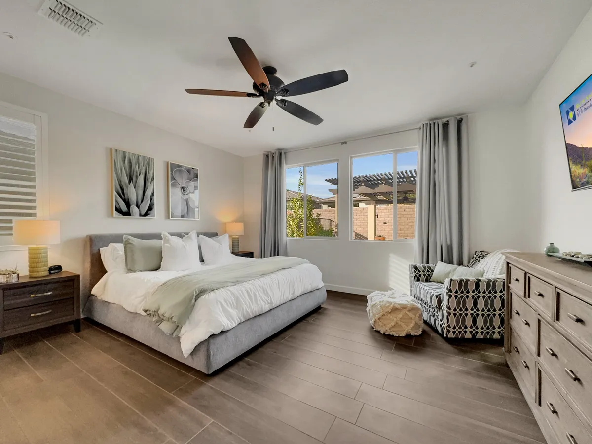 A spacious bedroom with a large bed adorned with decorative pillows, windows allowing in natural light, a cozy armchair for reading or relaxing, and tasteful wall art. The room exudes a sense of calm and comfort, ideal for unwinding at any time of day.