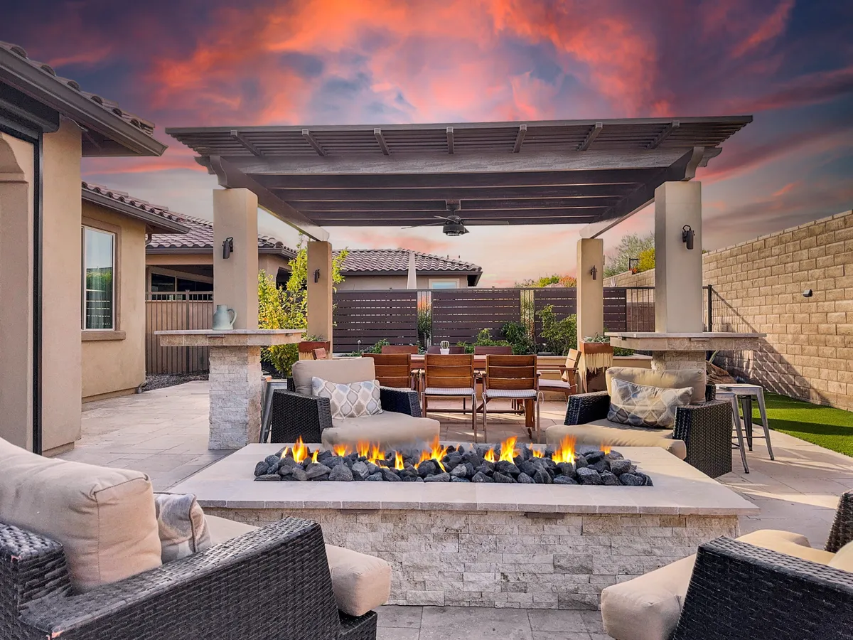 A beautifully designed outdoor living space featuring a cozy fire pit surrounded by plush seating, an outdoor dining table set for meals under a pergola, and a lush landscaped garden. The scene is set during the evening, with soft lighting creating a relaxed, intimate atmosphere ideal for gatherings or quiet relaxation.