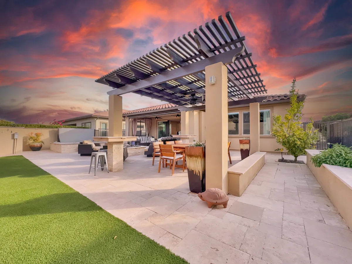 A beautifully designed exterior space featuring a pergola, comfortable outdoor seating, stone accents, lush greenery, and a stunning colorful sunset in the background. The scene is ideal for dining, social gatherings, or quiet moments of relaxation.